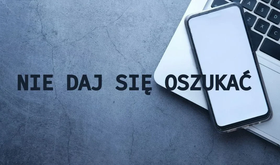 Smartfon z białym ekranem leży na klawiaturze laptopa na szarym betonowym tle, obok czarny napis ostrzegawczy.
