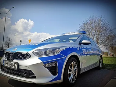 Policyjny radiowóz marki Kia z napisem „Policja” stoi zaparkowany na brukowanej nawierzchni przy drodze w słoneczny dzień.