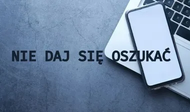 Smartfon z białym ekranem leży na klawiaturze laptopa na szarym betonowym tle, obok czarny napis ostrzegawczy.