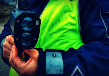 Funkcjonariusz w granatowej kurtce i żółtej kamizelce odblaskowej trzyma w dłoniach ręczny miernik prędkości laserowy.