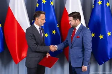 Dwaj mężczyźni w garniturach witają się uściskiem dłoni przed flagami Polski i Unii Europejskiej, jeden z nich trzyma czerwoną teczkę.
