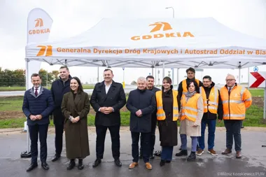Grupa osób, w tym urzędnicy i pracownicy drogowi w kamizelkach odblaskowych, pozuje pod namiotem GDDKiA podczas wydarzenia prasowego przy drodze.